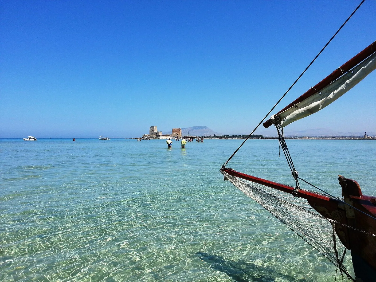 Acqua bassa e trasparente dello Stagnone a San Teodoro, con la prua di una barca di legno in primo piano e bagnanti in lontananza